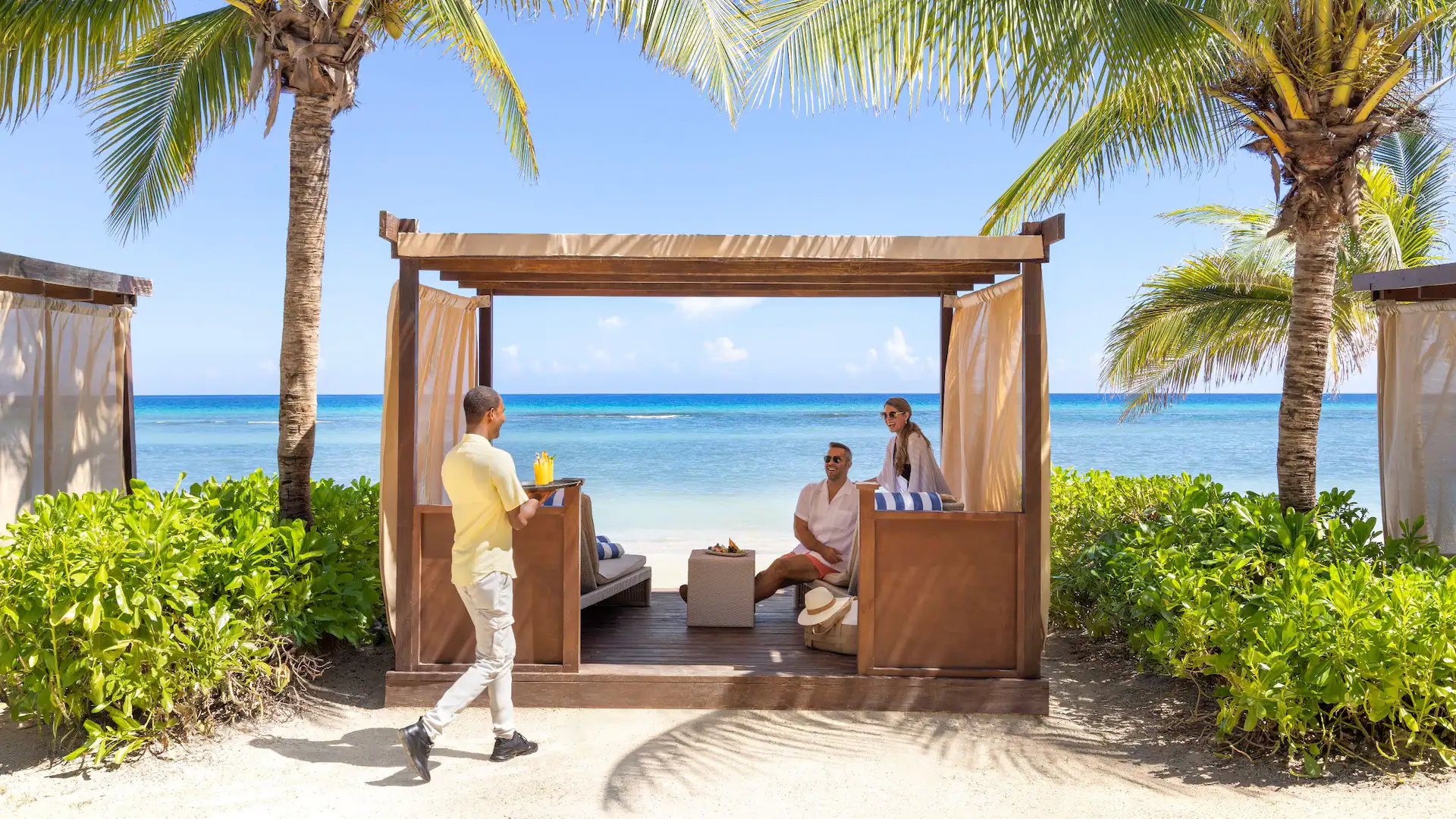 Couple relaxing in a beachfront cabana with personalized service at Dreams Rose Hall Resort & Spa in Jamaica.